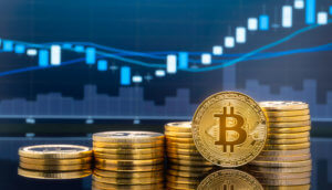 Eine aufgerichtete Bitcoin-Münze steht vor einigen aufgetürmten Bitcoin-Stapeln vor einer Chart.