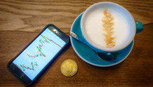 Smartphone das Charts zeigt, neben einer goldenen Bitcoin-Münze und einer Kaffeetasse mit Latteart auf einem braunen Holztisch