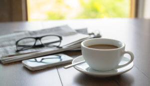 Tasse Kaffe auf dem Tisch mit einem Fenster im Hintergrund. Neben der Tase ein Smartphone und eine Zeitung, auf der eine zugeklappte Brille liegt