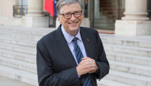 bill gates steht im anzug mit blau schwarzer krawatte und grauem haar vor einem gebäude, von dem man die treppenstufen sieht und lächelt