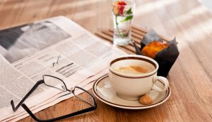 Eine Zeitung, eine Brille, eine Kaffeetasse samt Untertasse, ein Muffin und eine Blume stehen auf einem Holztisch.