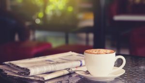 Eine Zeitung lieg neben einer Tasse Kaffee auf einem Steintisch. Dahinter fällt Licht durch ein Fenster in den Raum.